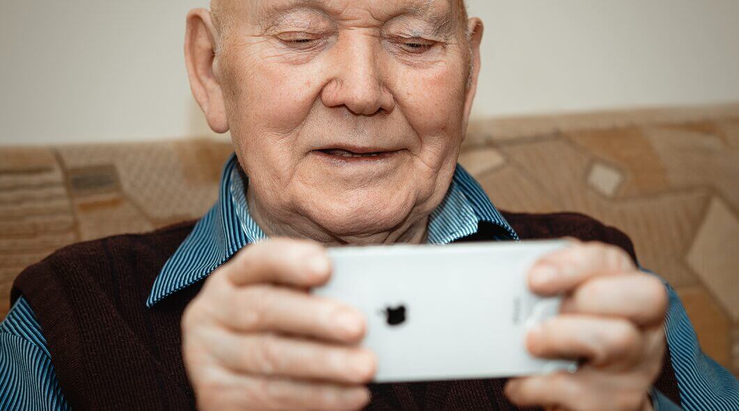 An old light skinned man sitting on a sofa is looking at a smartphone which is turned on its side