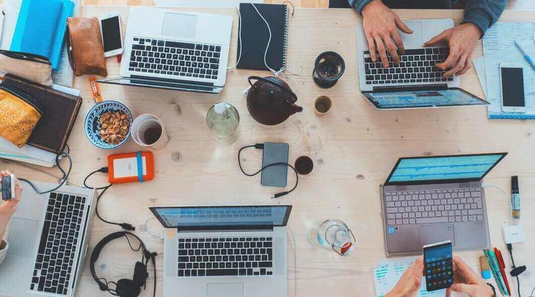 Bird's-eye view of a work desk cropped in so you can see two sets of hands using laptops and smartphones, other electrical items are strewn across the table surface