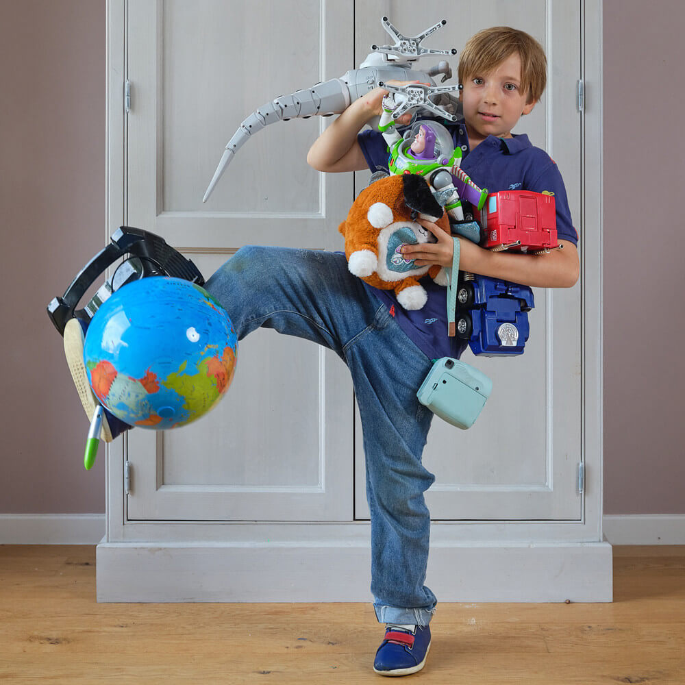a light skinned boy holding lots of toys in his arms and balancing some on his legs as well