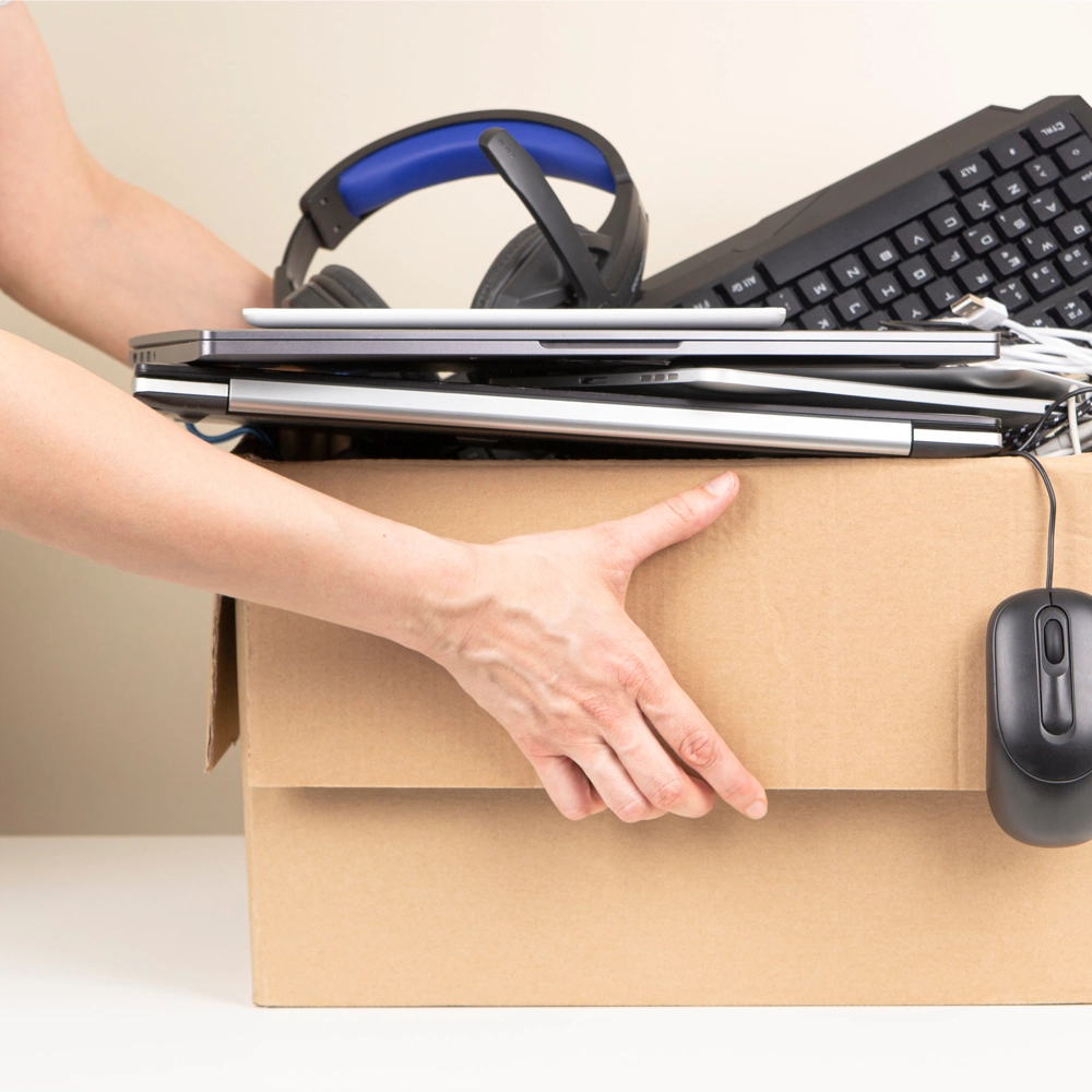 Light skinned person holding a cardboard box overflowing with electrical items such as headphones, a keyboard, laptops and a mouse.