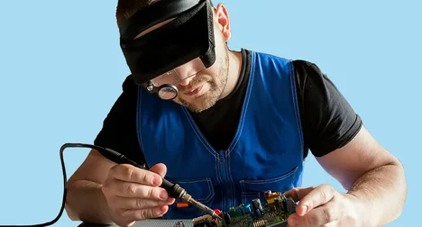 Light skinned man fixing a circuit board against a bright blue backdrop, he's wearing a magnifying visor and holding a soldering iron in one hand and the circuit board tilted in the other.