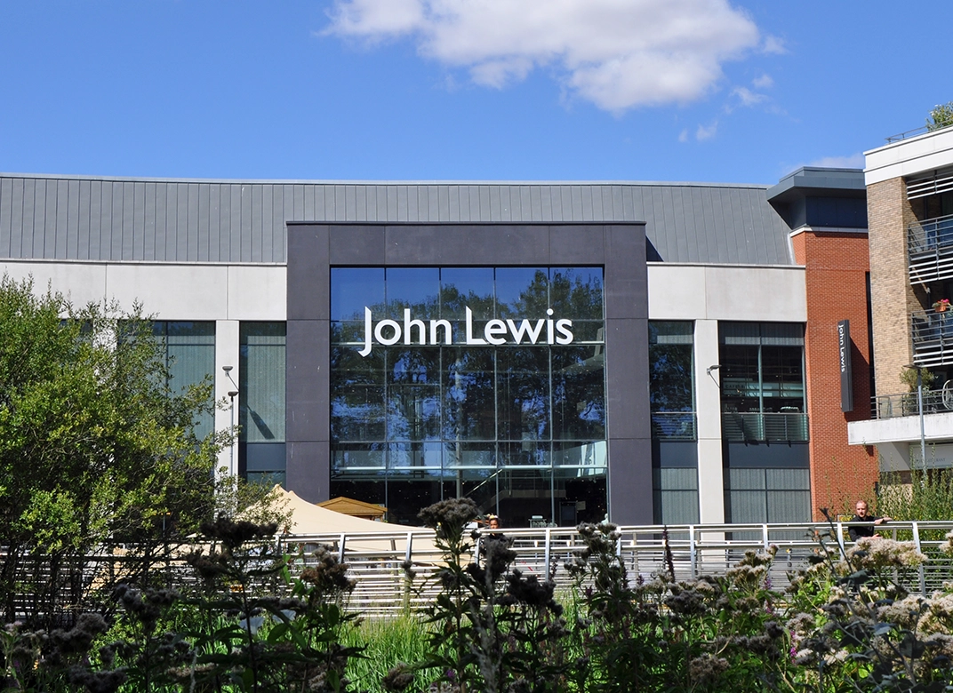 Entrance of a John Lewis store with shiny glass windows reflecting the blue sky and trees surrounding the building.