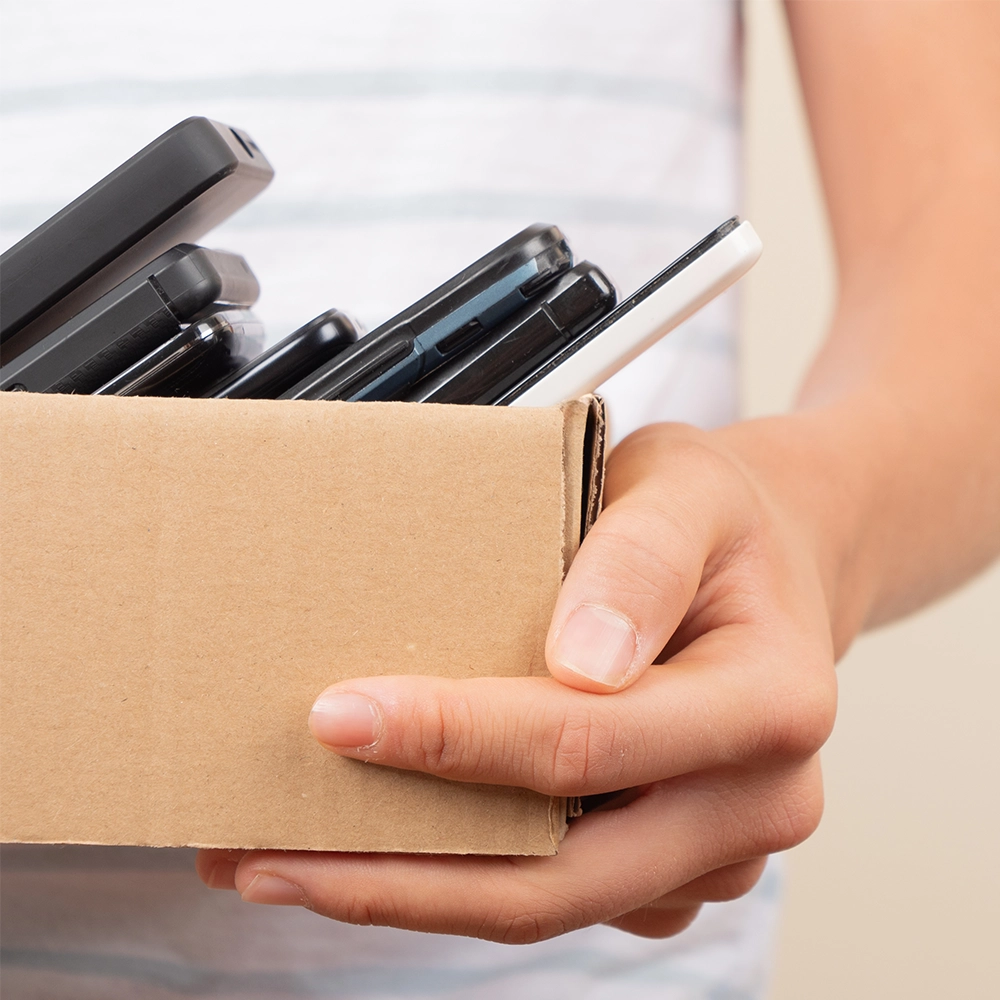 Close up of a light skinned person holding a small cardboard box full of stacked mobile phones.