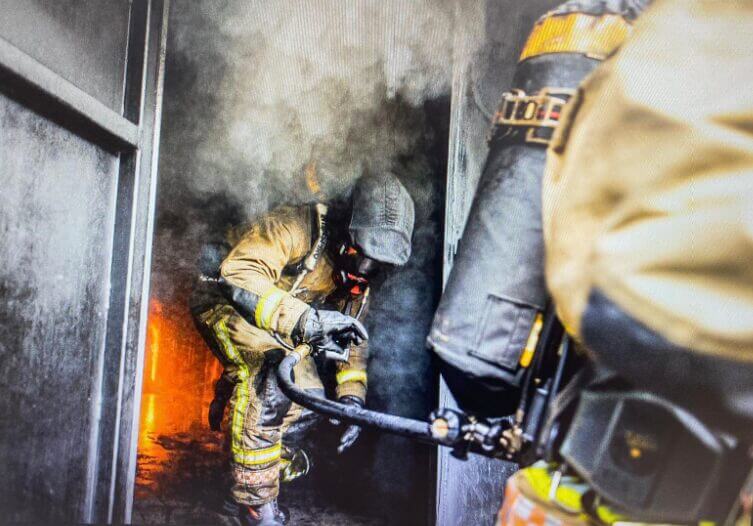 fire officer wearing yellow uniform wearing black helment and carrying hose, escaping from burning building with fire burning in background