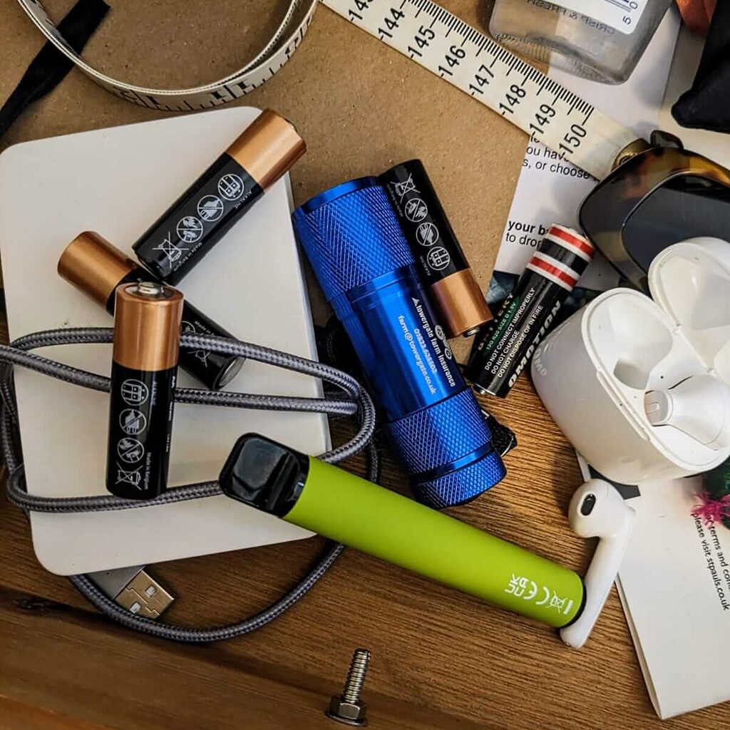 drawer of old batteries, old green vape, white phone battery pack with grey cable wrapped around, blue torch and white air pod headphones