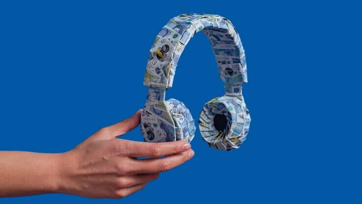 hand holding headphones wrapped in five pound notes against a blue background